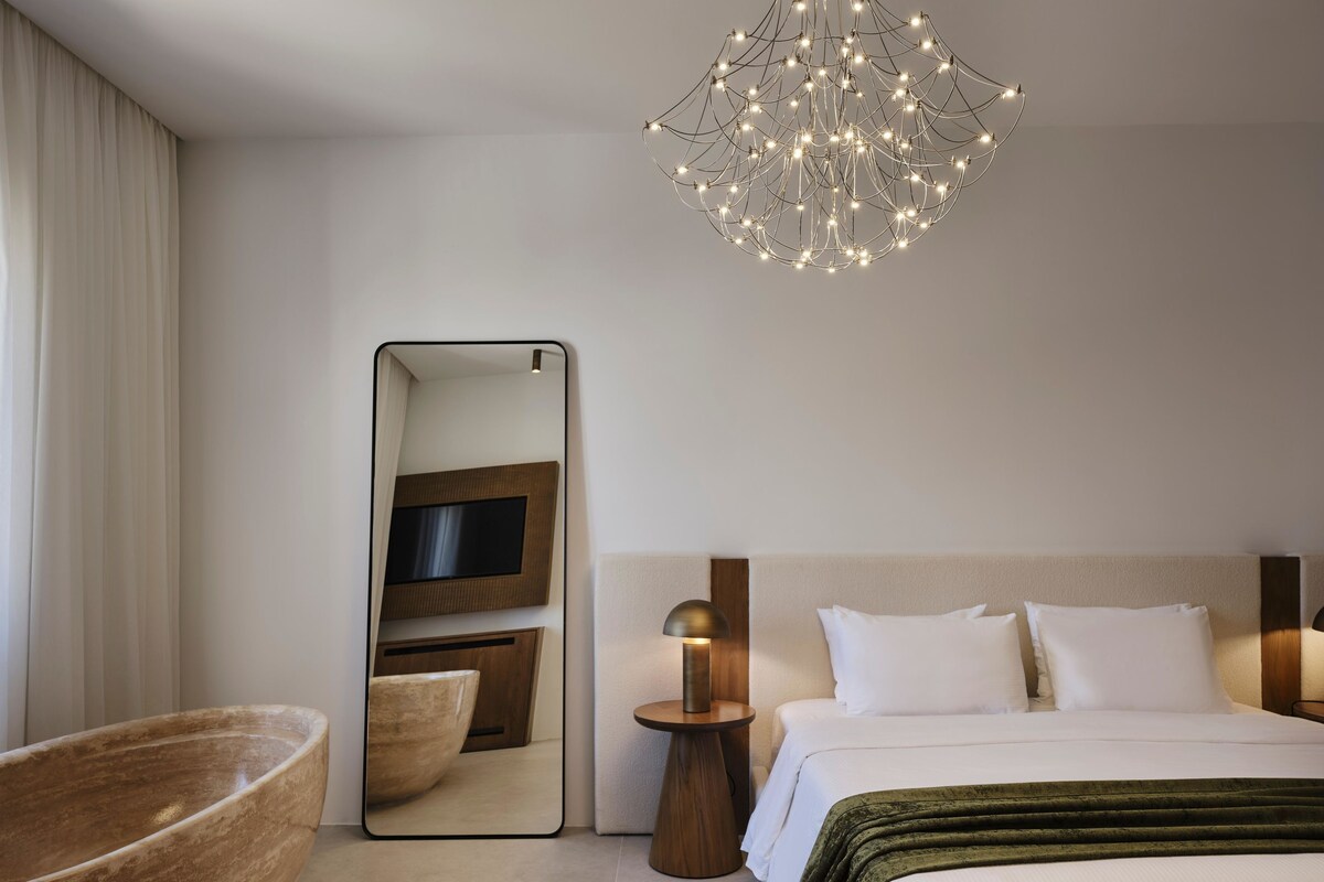 Una habitación de hotel moderna con una cama grande, una mesita de madera, un espejo y una lámpara de araña original.