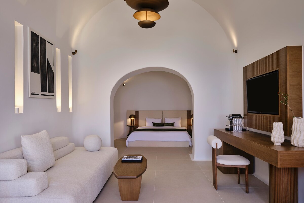 Una habitación de hotel moderna con sofá, una mesa de centro de madera, un escritorio con silla, televisión montada en la pared y una cama grande con cabecera.