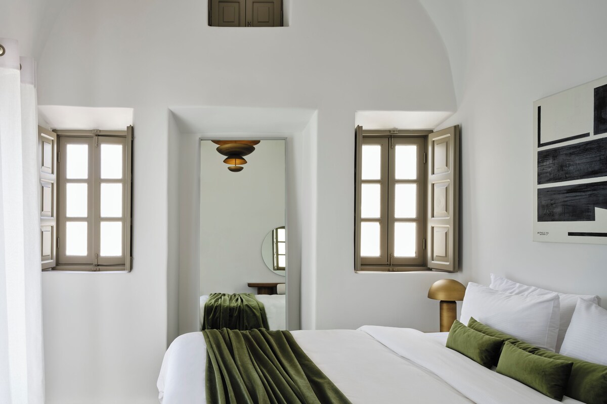 Un dormitorio con una cama grande, dos ventanas con contraventanas abiertas, un espejo y un cuadro en la pared.