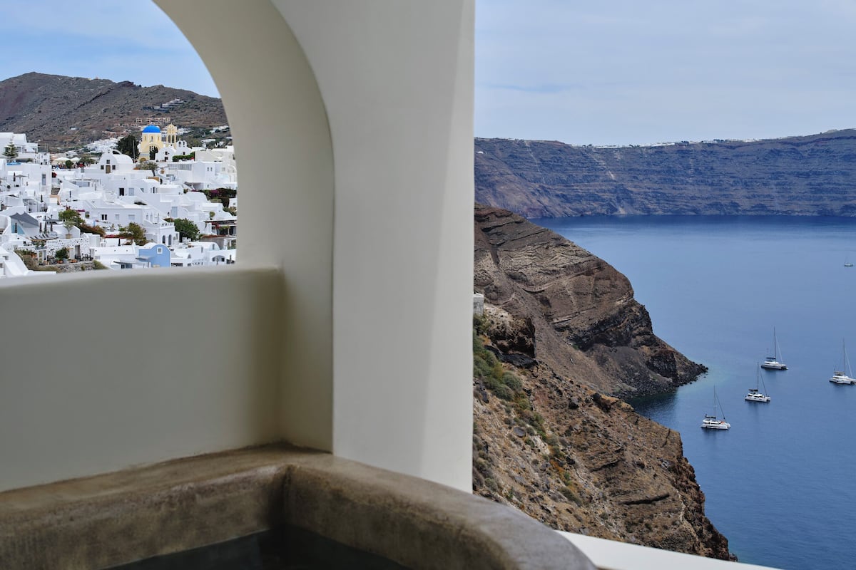 A view from a balcony with a stone bathtub, overlooking a coastal town with white buildings and boats on the water.