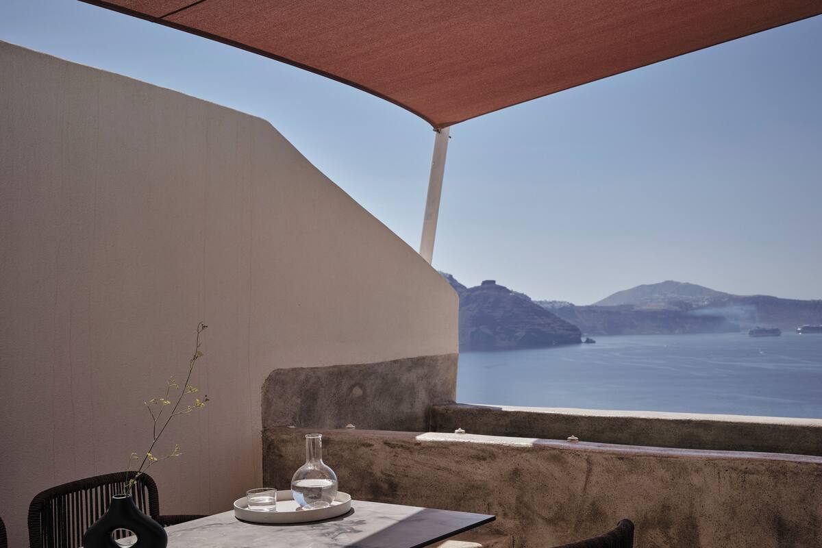 A terrace with a table, chairs, a vase with a branch, and a bottle. The terrace overlooks a body of water and distant landforms.