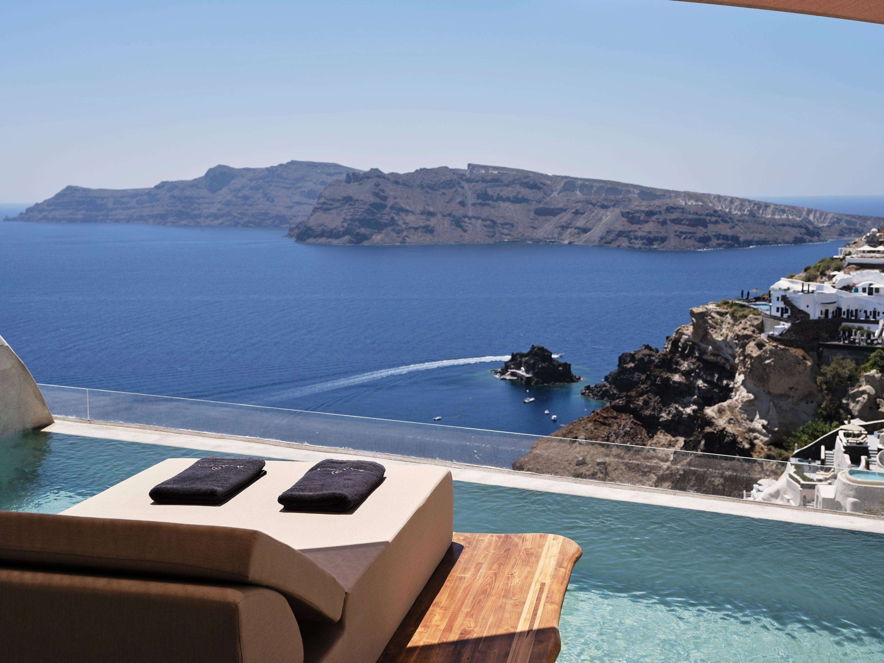 A poolside view with a wooden deck, lounge chairs, and a view of the sea and rocky coastline.