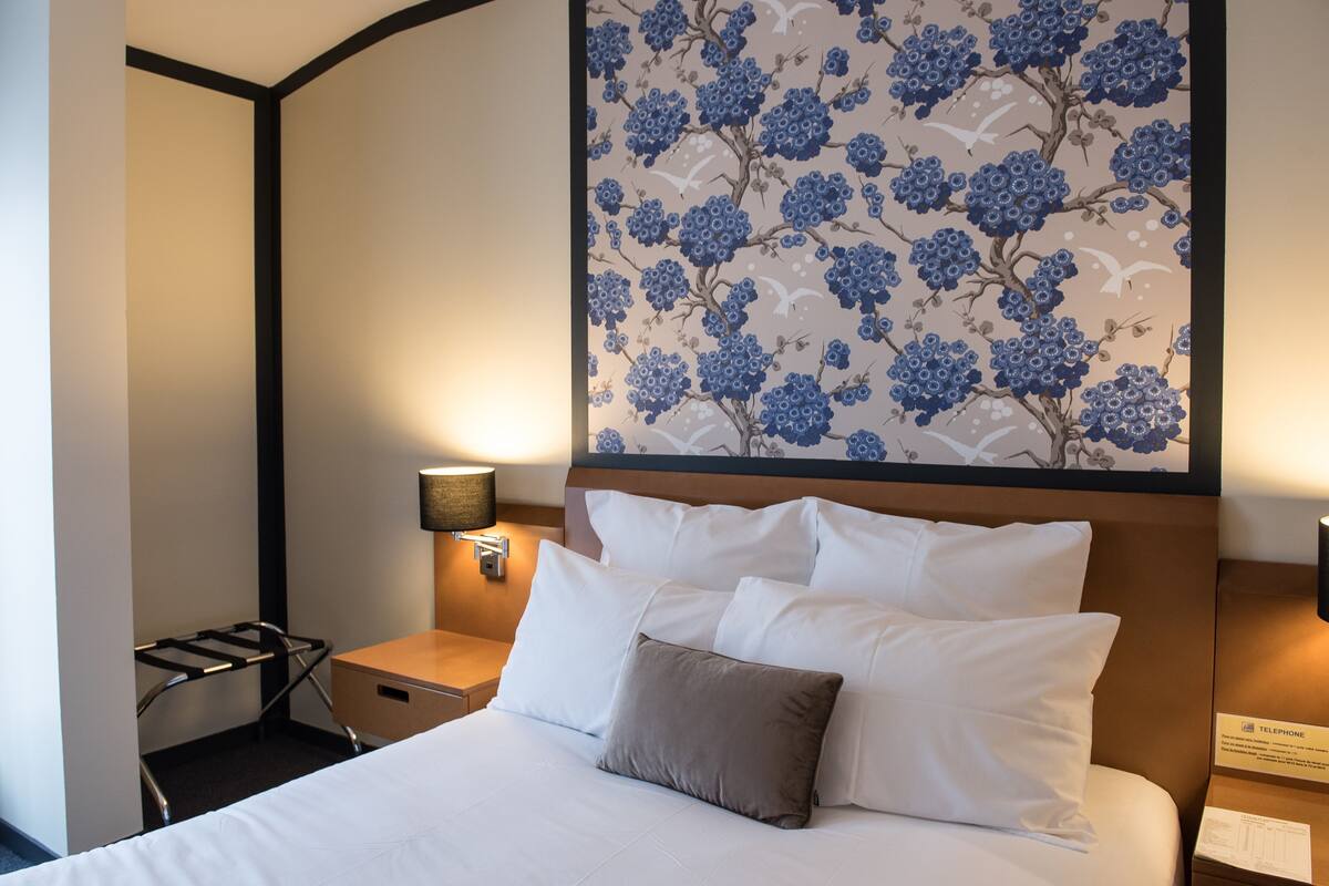 Una habitación de hotel con una cama bien hecha, una mesita de noche con una lámpara y una pared con estampado floral.