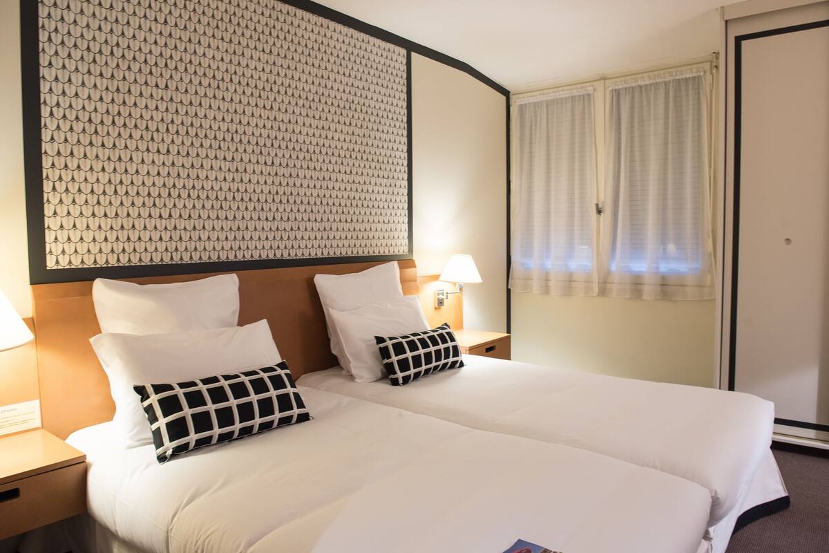 Una habitación de hotel con una cama grande, dos mesitas de noche con lámparas, un cabecero con diseño texturizado y una ventana con cortinas.