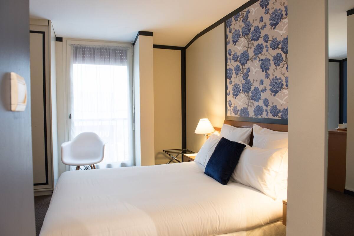 Habitación de hotel con una cama grande, una silla, una mesita de noche con lámpara y una ventana con cortinas.