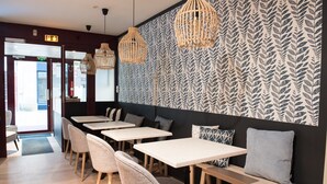 Interior de restaurante con mesas y sillas blancas, pared con diseño y lámparas colgantes de mimbre.