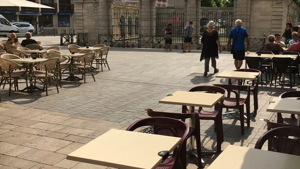 Un café al aire libre con mesas y sillas, un arco de piedra y un edificio con un letrero al fondo.