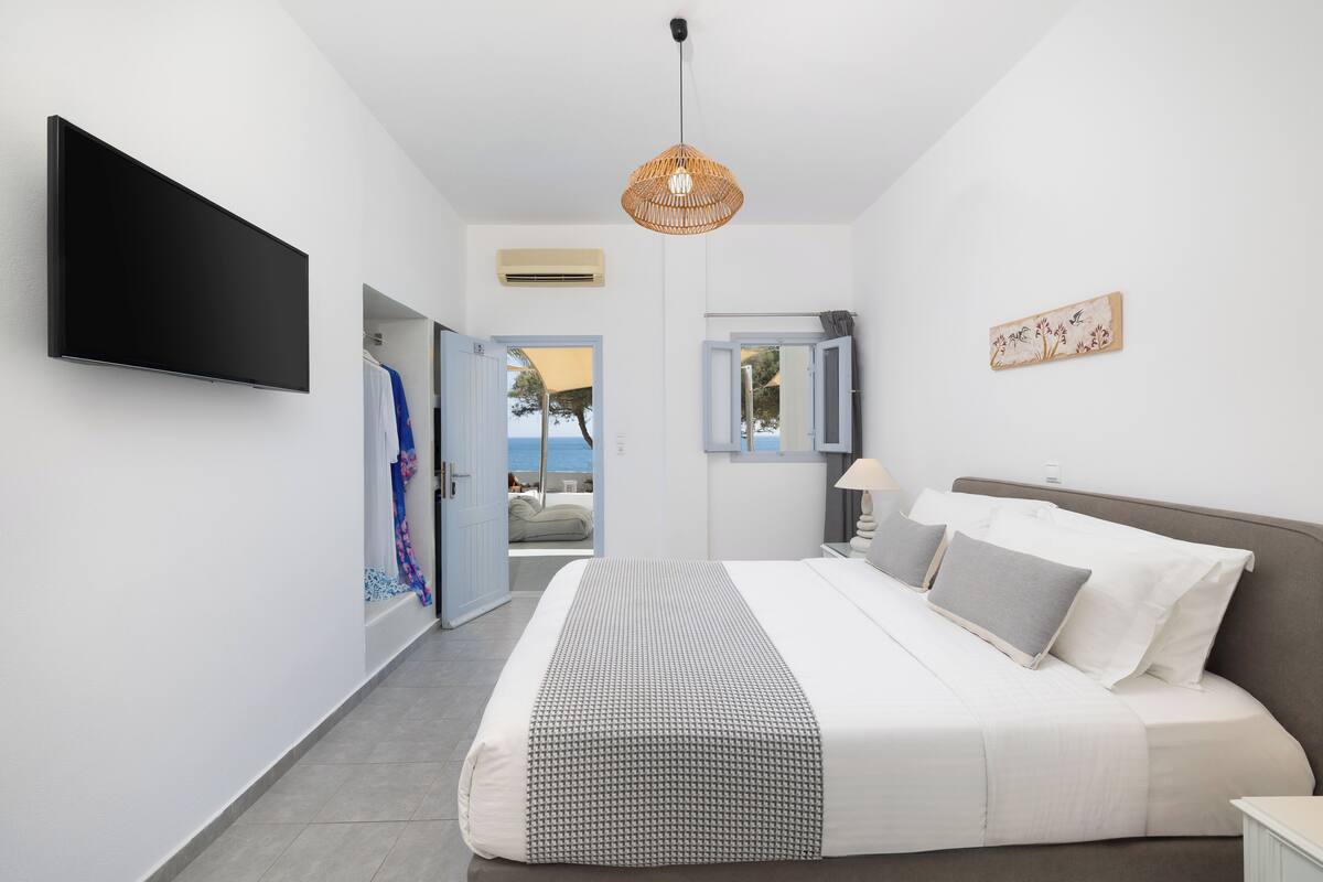 A modern bedroom with a large bed, a flat-screen TV, a rattan ceiling light, and a view of the sea through an open door.