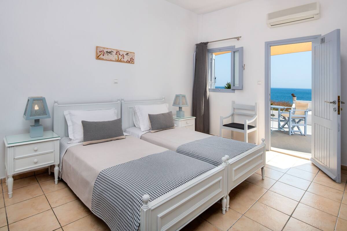 A bedroom with two beds, a small table, and a chair. There is a window with a view of the sea.