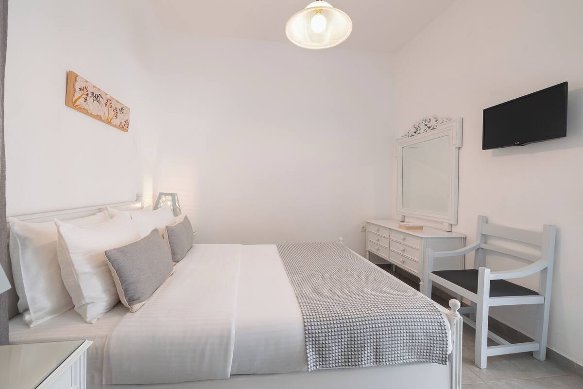 A bedroom with a bed, bedside table, chair, dresser, mirror, and a flat-screen TV.