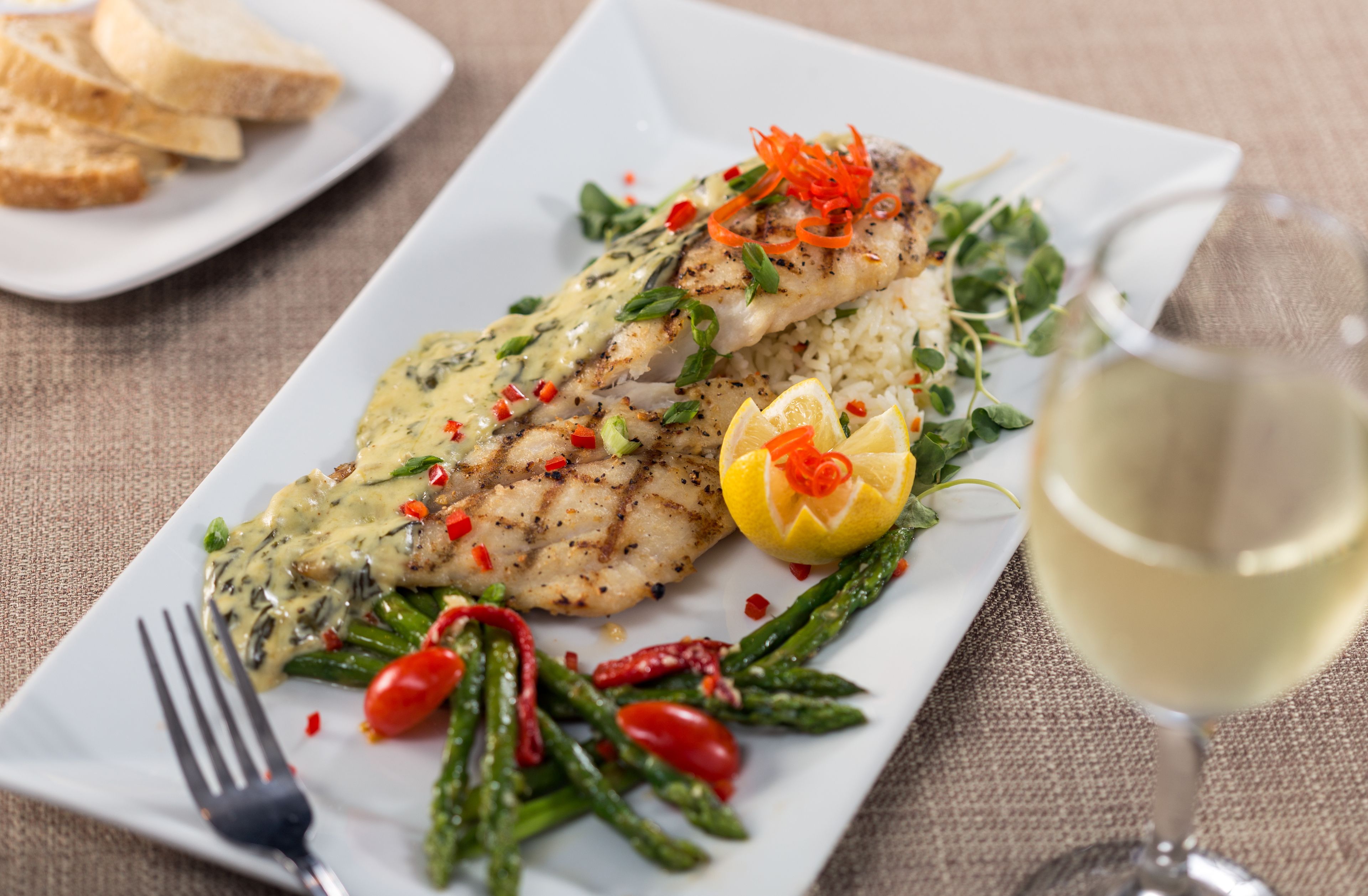 A plate of grilled fish with a creamy sauce, garnished with herbs and vegetables, accompanied by a glass of white wine.