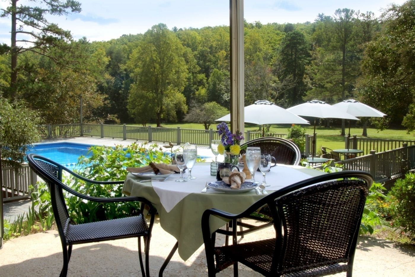An outdoor dining area with a table set for two, overlooking a swimming pool and a forested area.