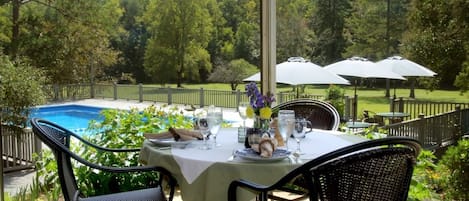 An outdoor dining area with a table set for two, overlooking a swimming pool and a forested area.