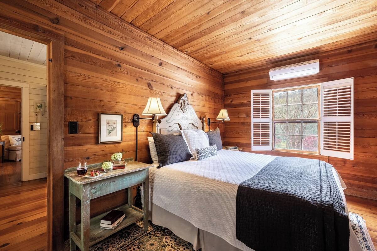 A wooden cabin bedroom with a bed, bedside lamps, a nightstand, and a window with shutters.
