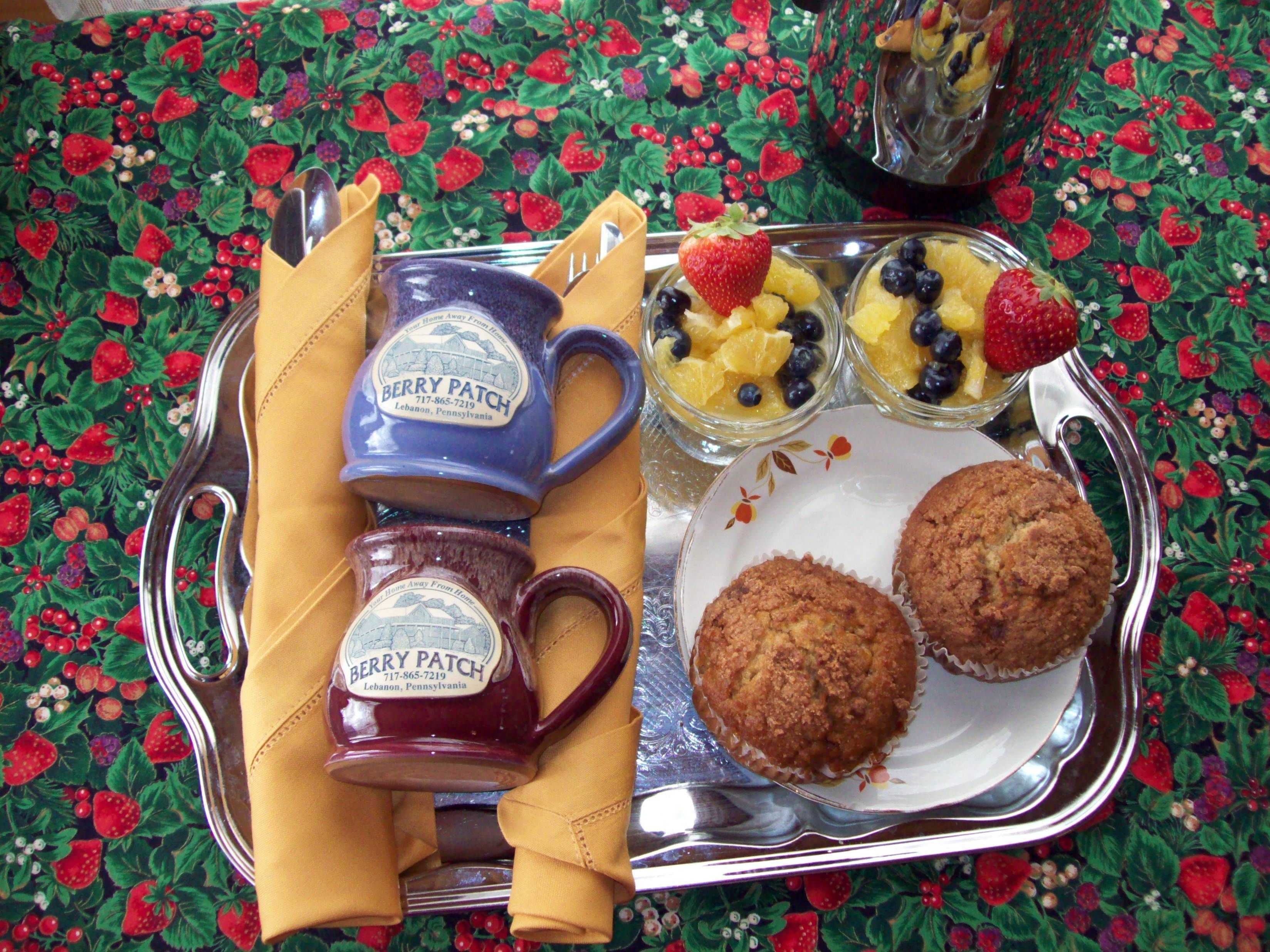 A tray with two mugs, a plate with muffins, and a bowl of fruit.