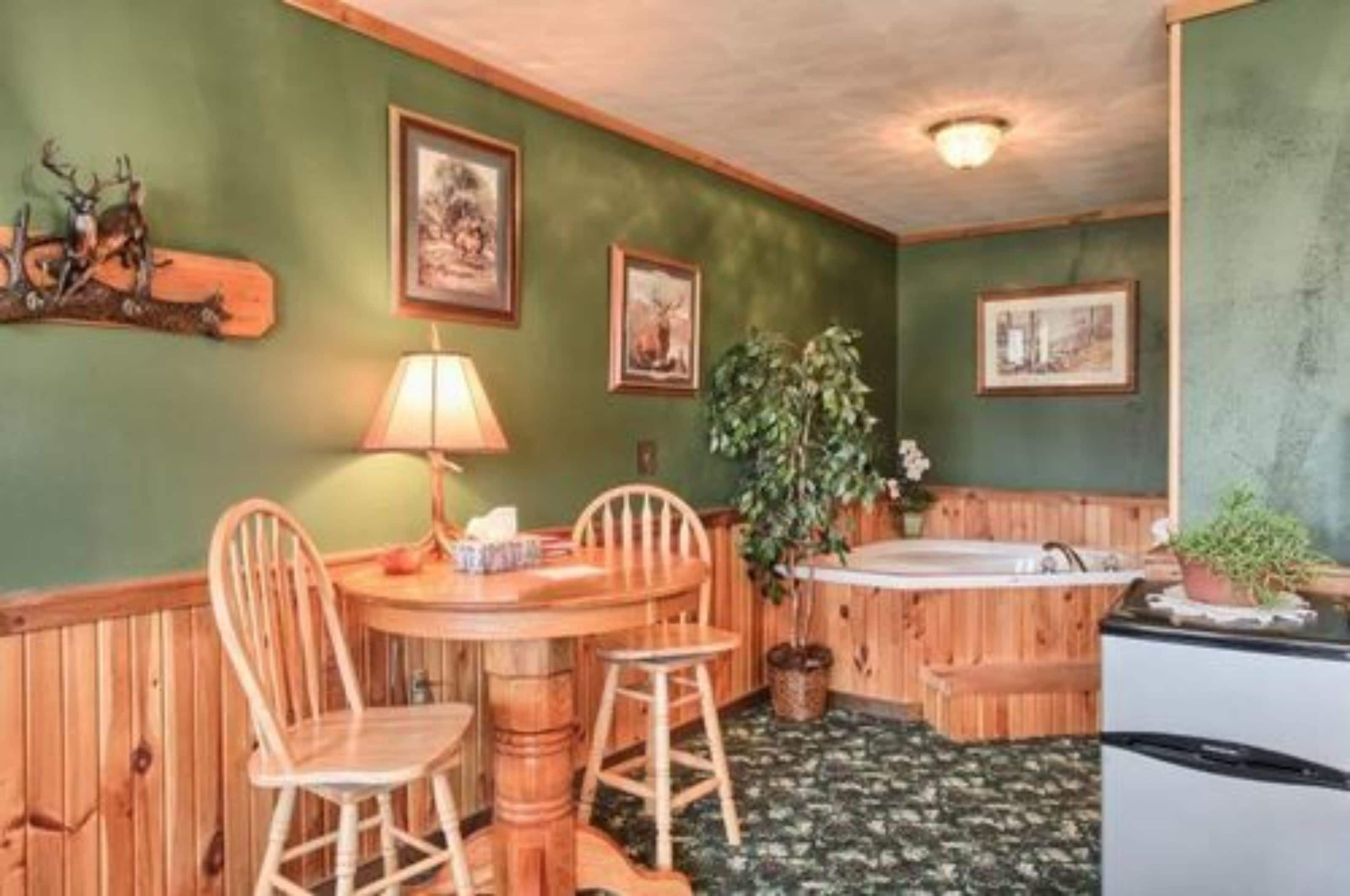 A room with a wooden table, two chairs, a lamp, a plant, a bathtub, and framed pictures on the wall.