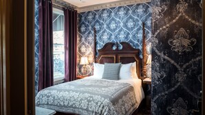 A bedroom with a four-poster bed, a nightstand with a lamp, and a patterned wallpaper.