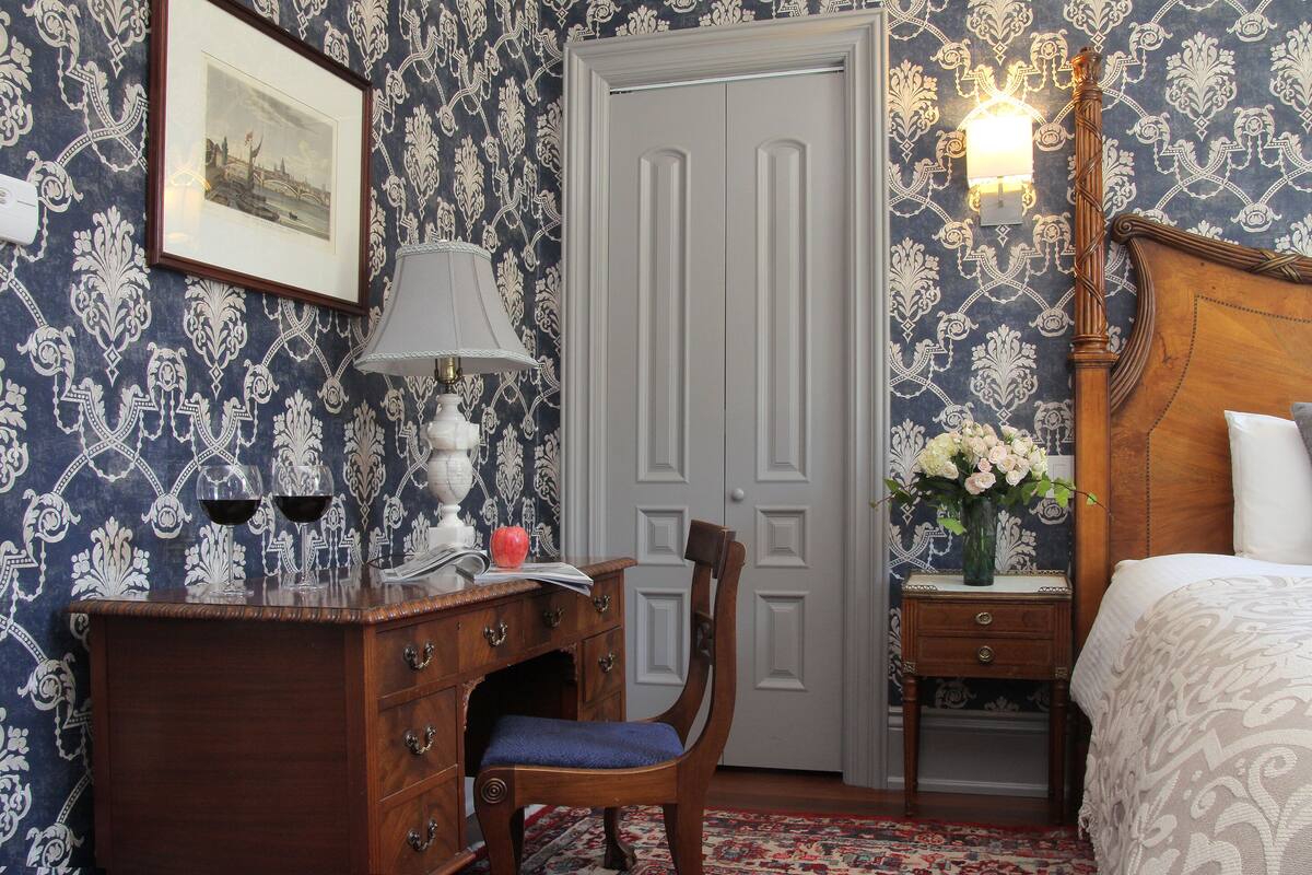 A room with a wooden dresser, a chair, a desk with a lamp, a bed with a patterned bedspread, and a door with a light fixture.