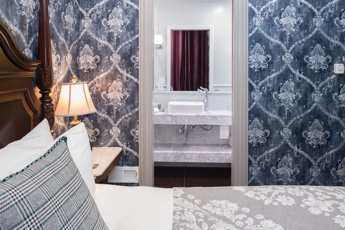 A bedroom with a bed, a nightstand with a lamp, a bathroom with a sink and mirror, and patterned wallpaper.