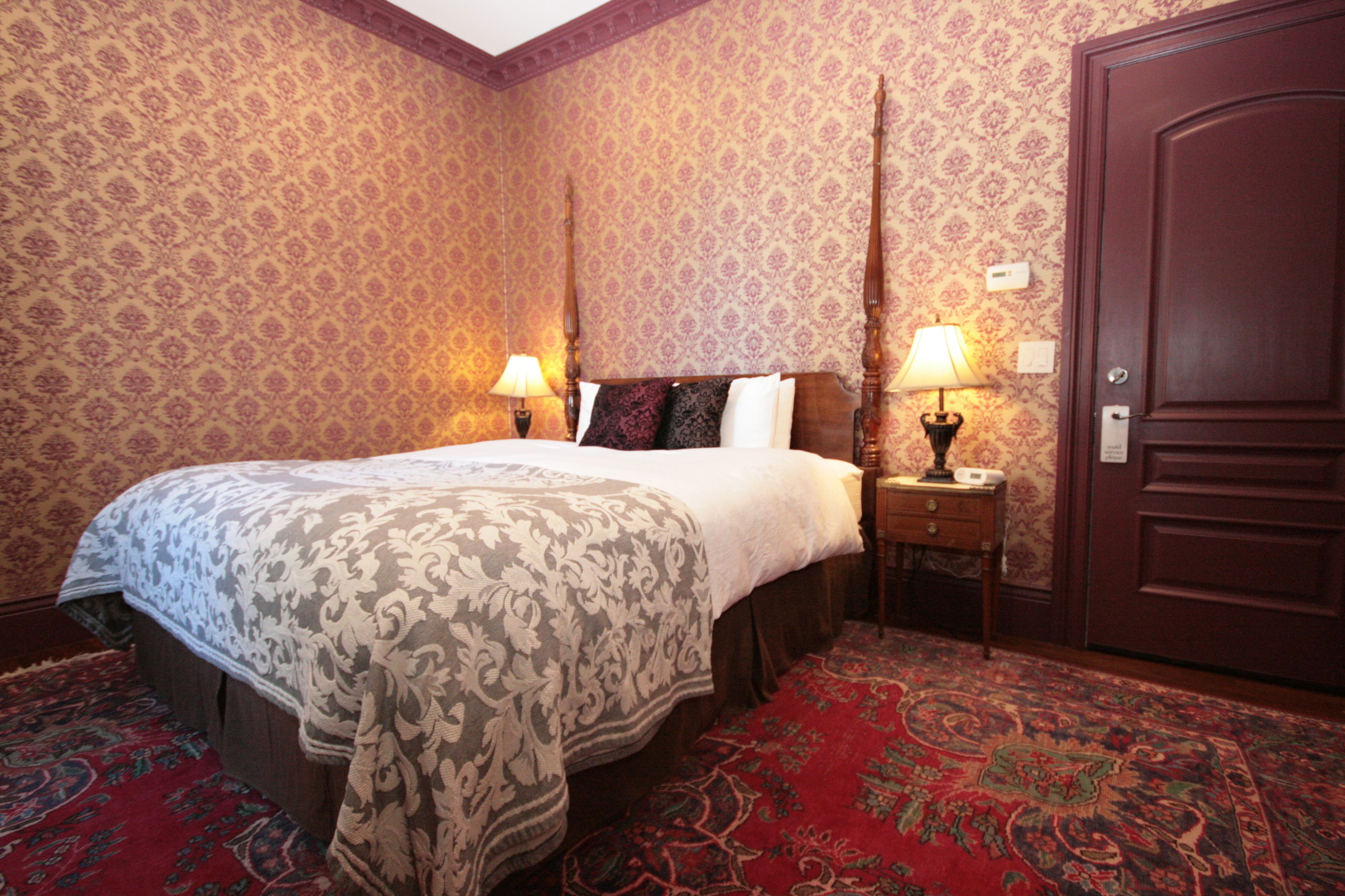 A hotel room with a large bed, two bedside tables with lamps, a wooden door, and patterned wallpaper.