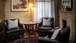 A cozy living room with two leather armchairs, a small round table, a lamp, a framed picture on the wall, and a brick fireplace.