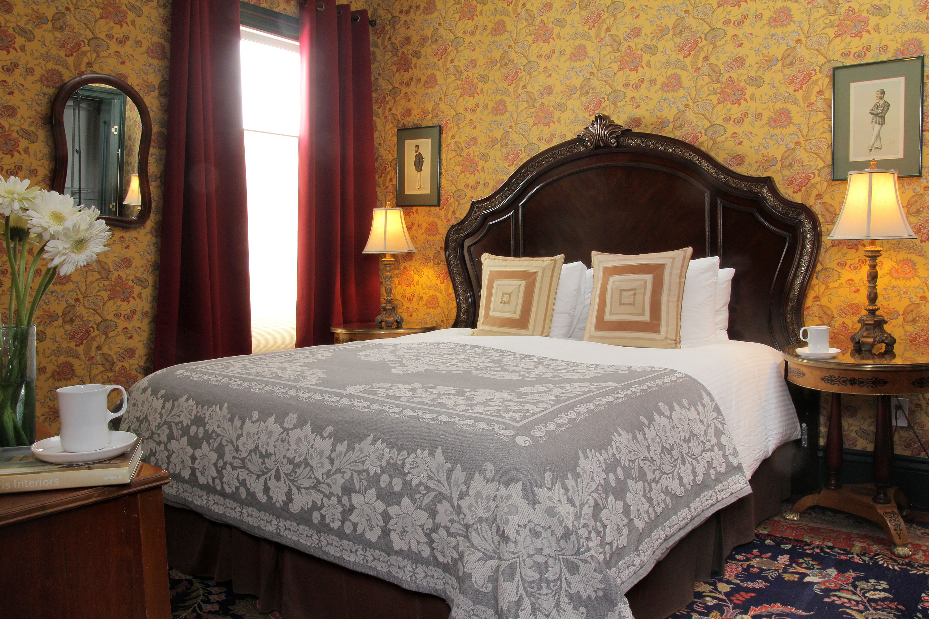 A hotel room with a large bed, floral wallpaper, a wooden headboard, two bedside lamps, a small table with a cup and saucer, and a vase with flowers.