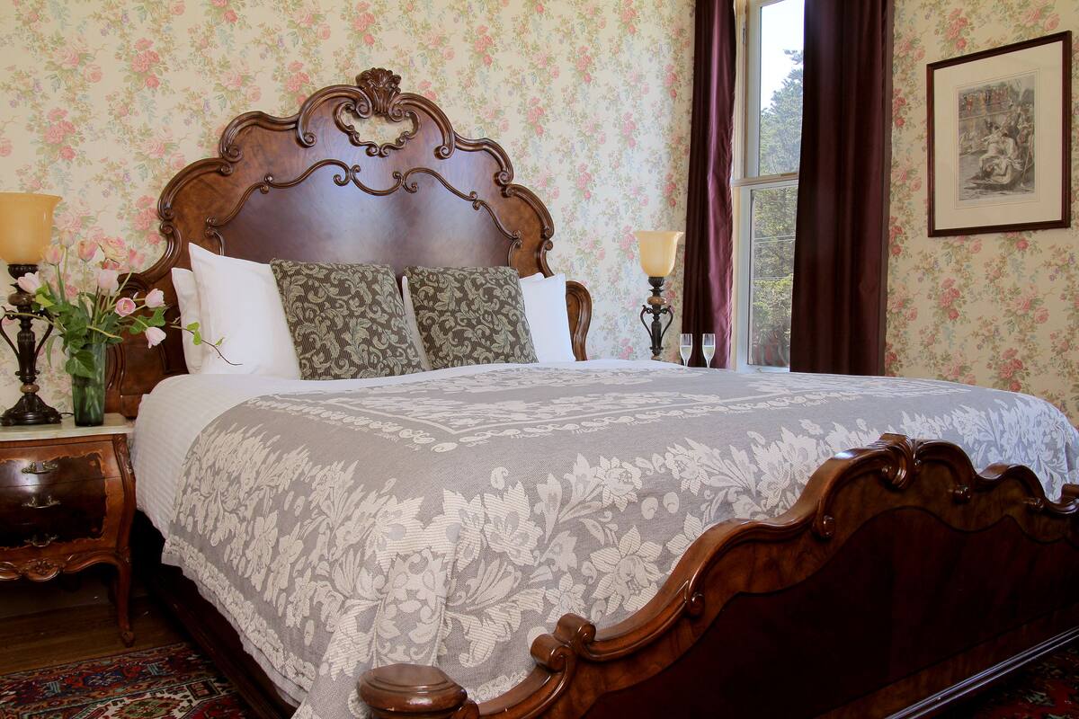 A classic wooden bed with a floral-patterned bedspread and matching pillows, set against a wallpapered wall with a framed picture.