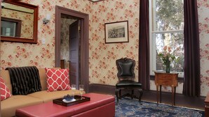 A room with floral wallpaper, a sofa with red and white cushions, a red ottoman, a wooden chair, a small table with a vase, and a large window with curtains.