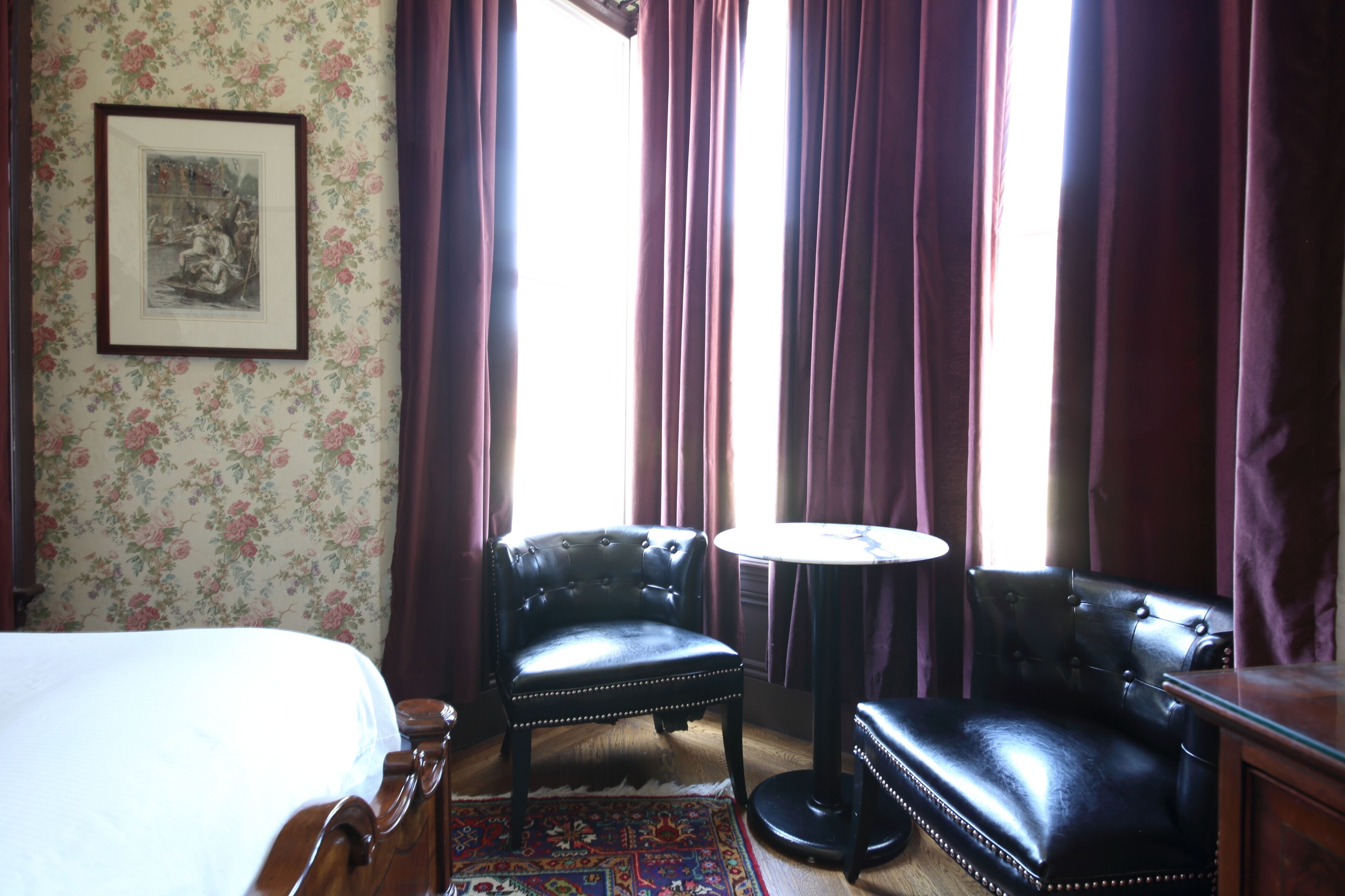 Una habitación con una cama, dos sillones de cuero, una mesita redonda y un papel tapiz floral.