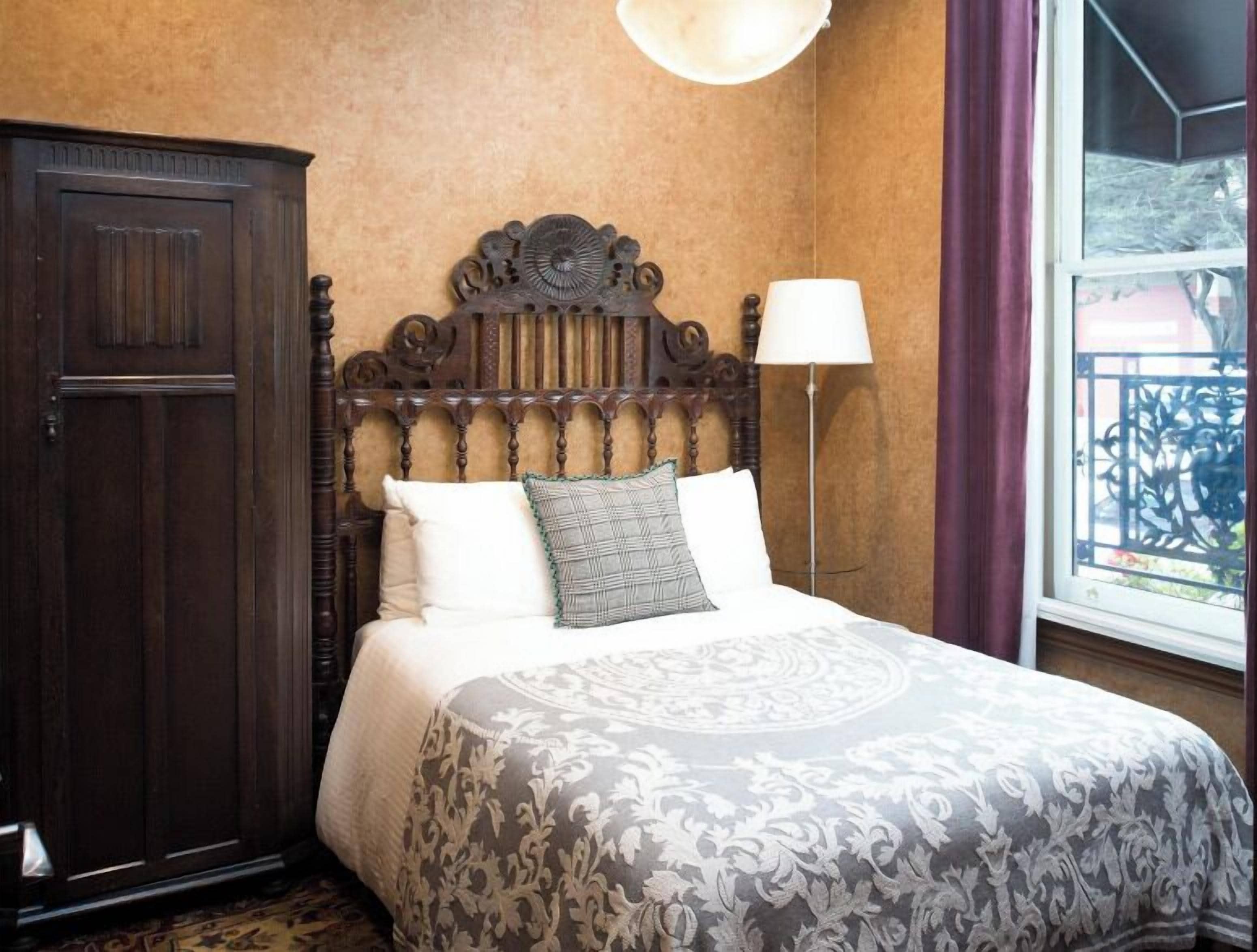A room with a large dark wooden wardrobe, a bed with a patterned bedspread, a floor lamp, and a window with a view of a balcony.