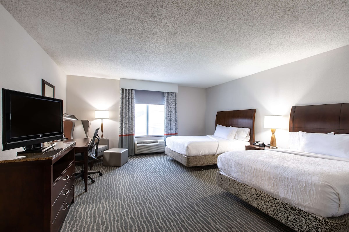 A hotel room with a flat-screen TV, two beds, a desk with a chair, a lamp, and a window with curtains.