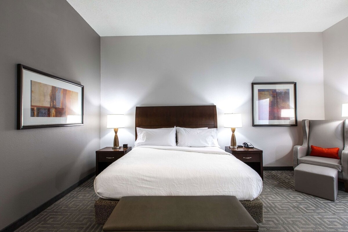 Een hotelkamer met een groot bed, twee nachtkastjes, een stoel, een bankje en twee ingelijste foto's aan de muur.