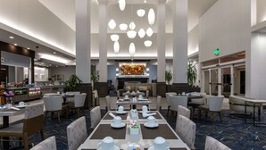 A hotel dining area with tables set for service, a central decorative wall, and a buffet area in the background.