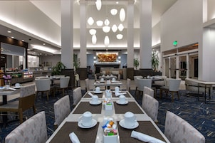 A hotel dining area with tables set for service, a central decorative wall, and a buffet area in the background.