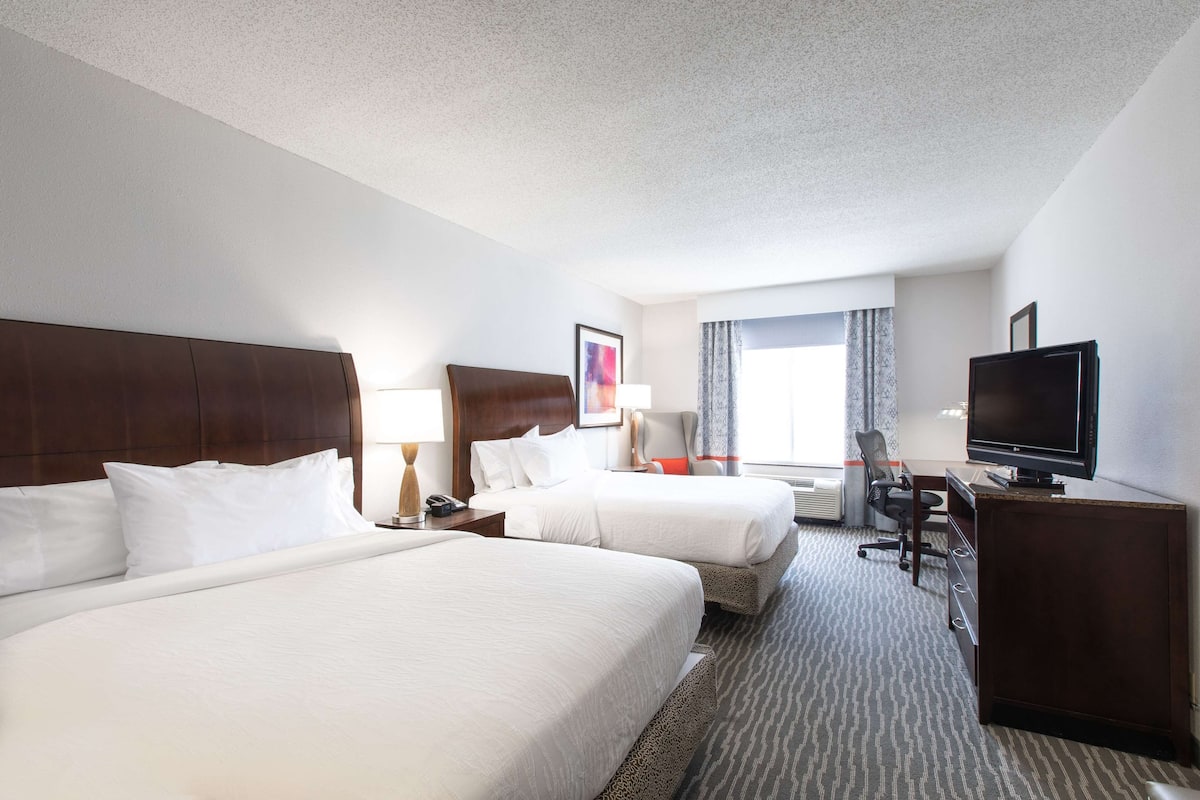 A hotel room with two beds, a desk, a chair, a television, and a window with curtains.