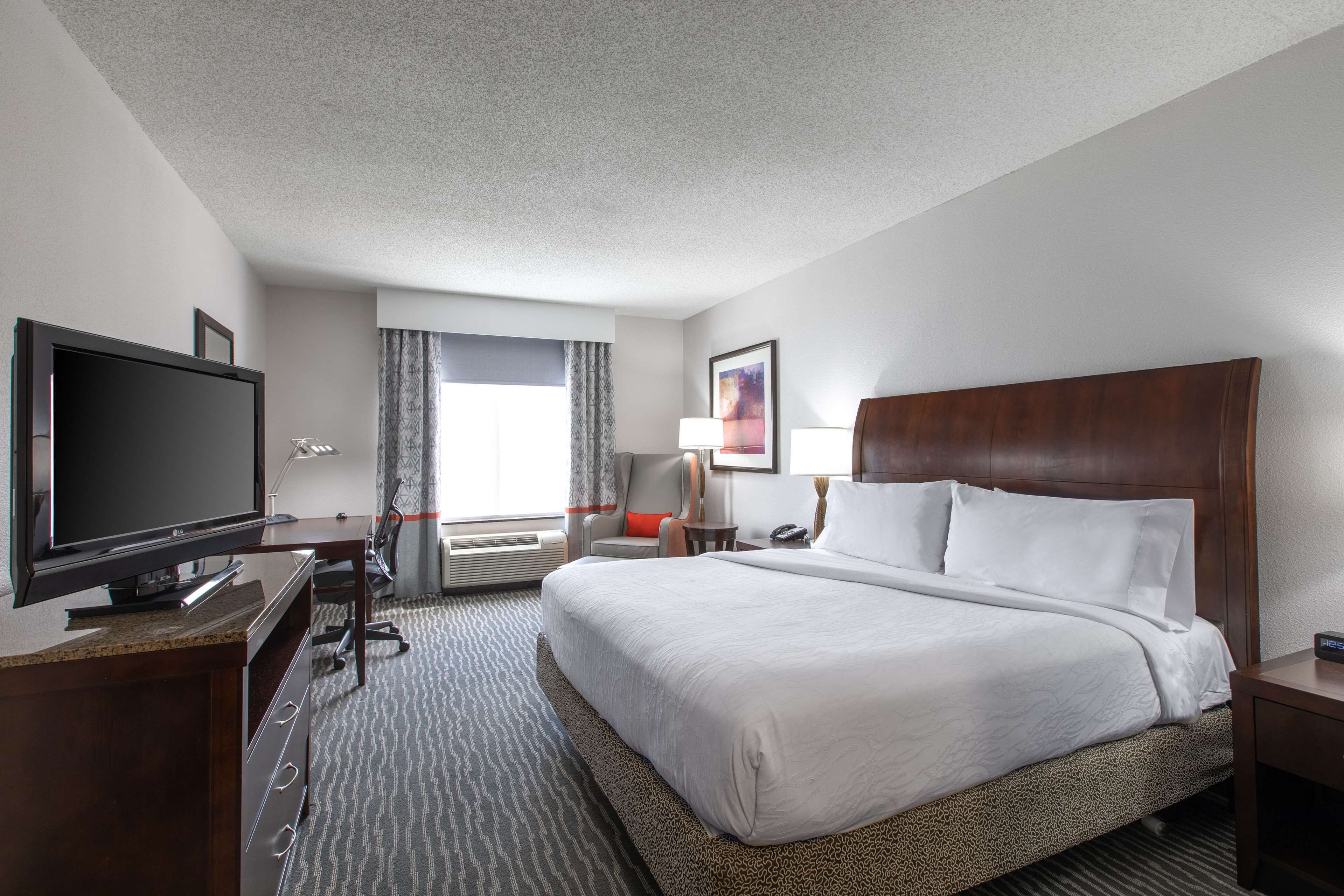 A hotel room with a large bed, a flat-screen TV, a desk with a computer, a chair, and a window with curtains.