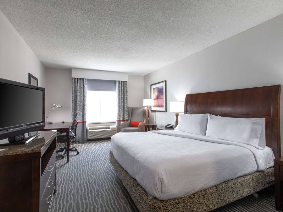 A hotel room with a large bed, a flat-screen TV, a desk with a computer, a chair, and a window with curtains.