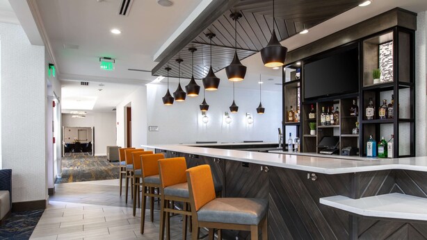 A modern bar area with a long counter, multiple bar stools, pendant lighting, and a shelf with bottles.