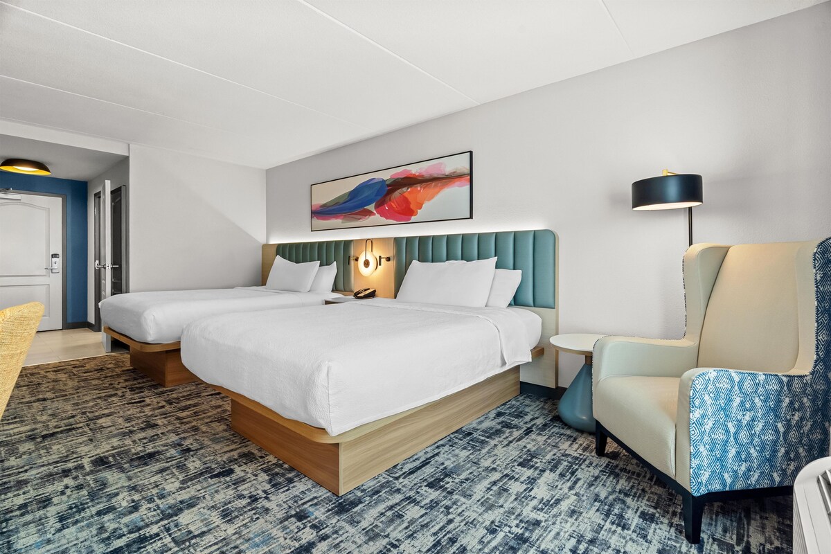 A hotel room with a large bed, a bedside table, a chair, a desk, and a painting on the wall.