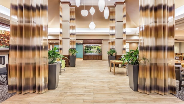 Een ruime lobby met een houten vloer, grote potplanten en een receptiebalie.