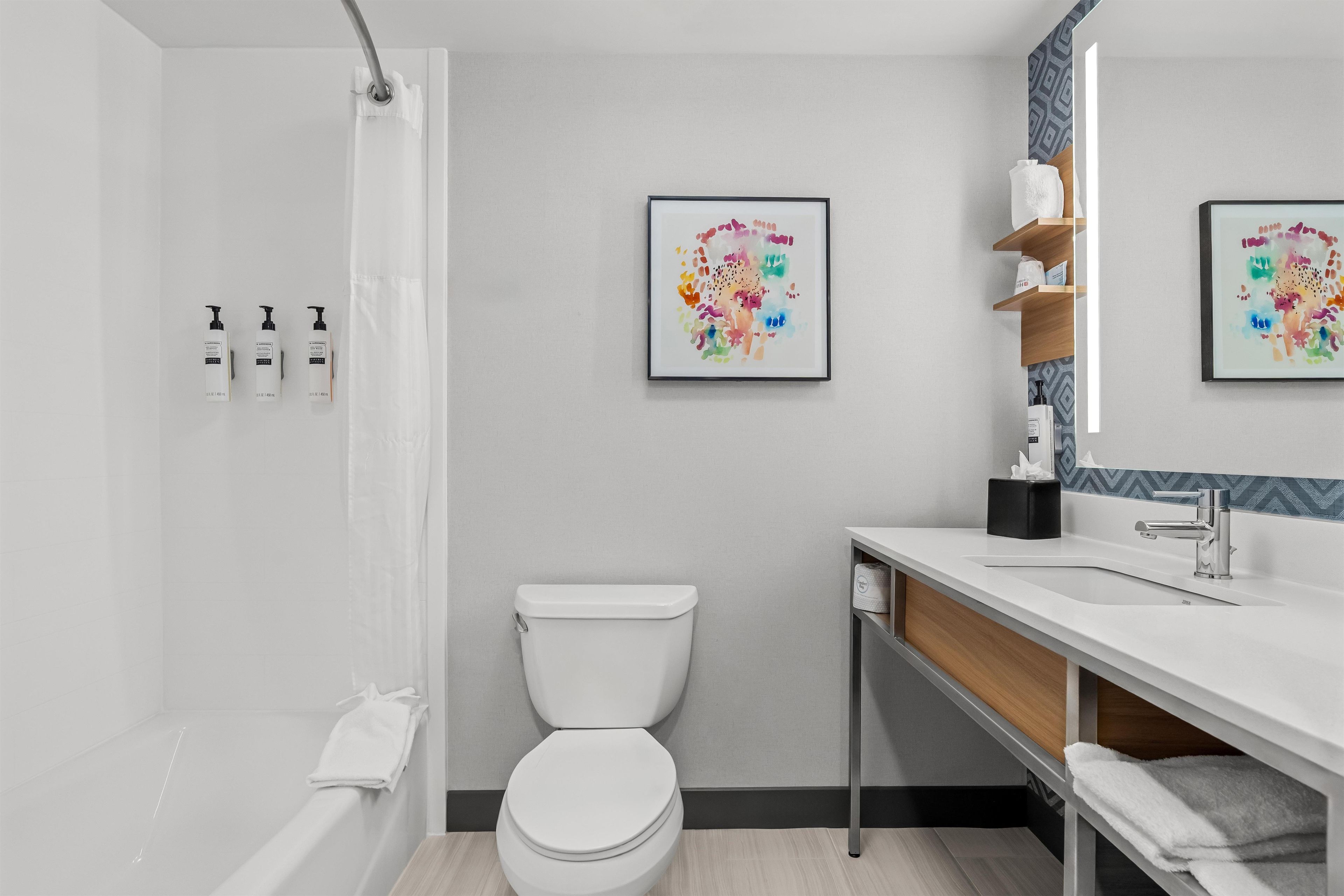 Un baño moderno con ducha, inodoro y lavamanos. Hay obras de arte enmarcadas en la pared, un perchero y un estante con artículos de aseo.