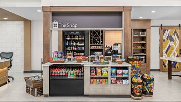 Quiosco de aperitivos y bebidas llamado 'The Shop', con secciones de productos fríos y regulares, diversas opciones de comida y un área de descanso con una silla y una mesa.