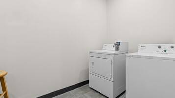 A laundry room with two white Whirlpool washers and dryers, a wooden stool, and grey tiled flooring.