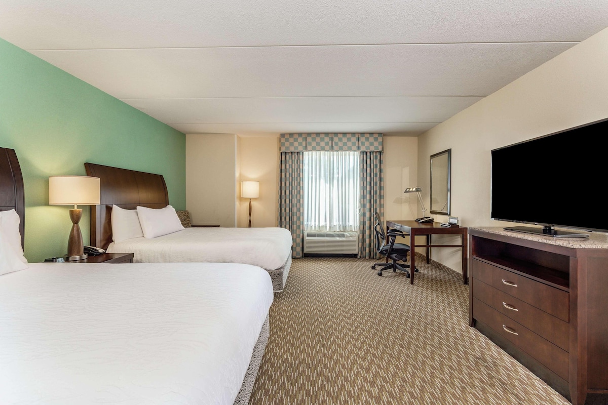 A hotel room with two beds, a desk, a television, and a window with curtains.