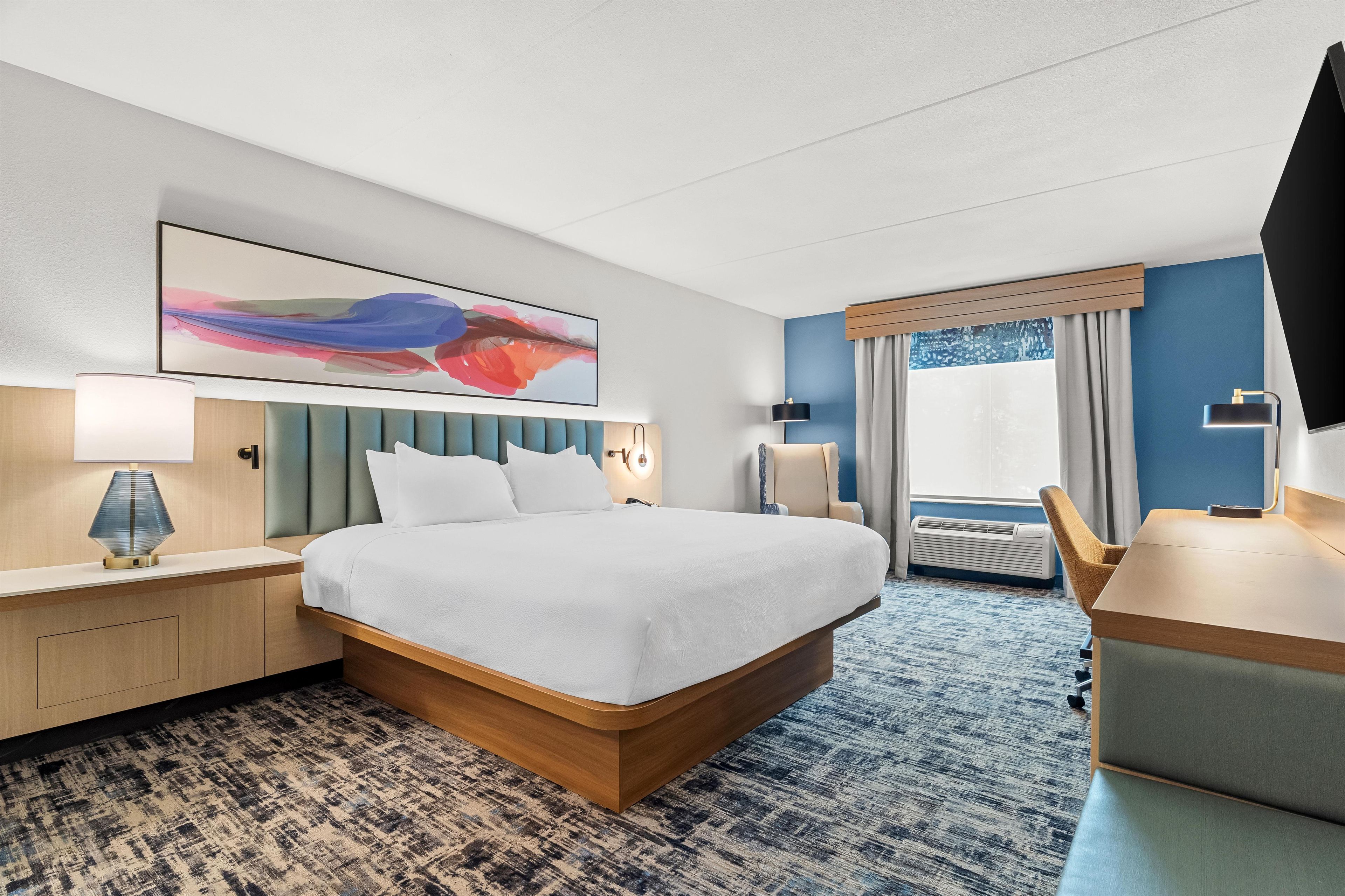 Une chambre d’hôtel avec un grand lit, un bureau, une chaise, une lampe de bureau, un téléviseur fixé au mur et une fenêtre avec des rideaux.