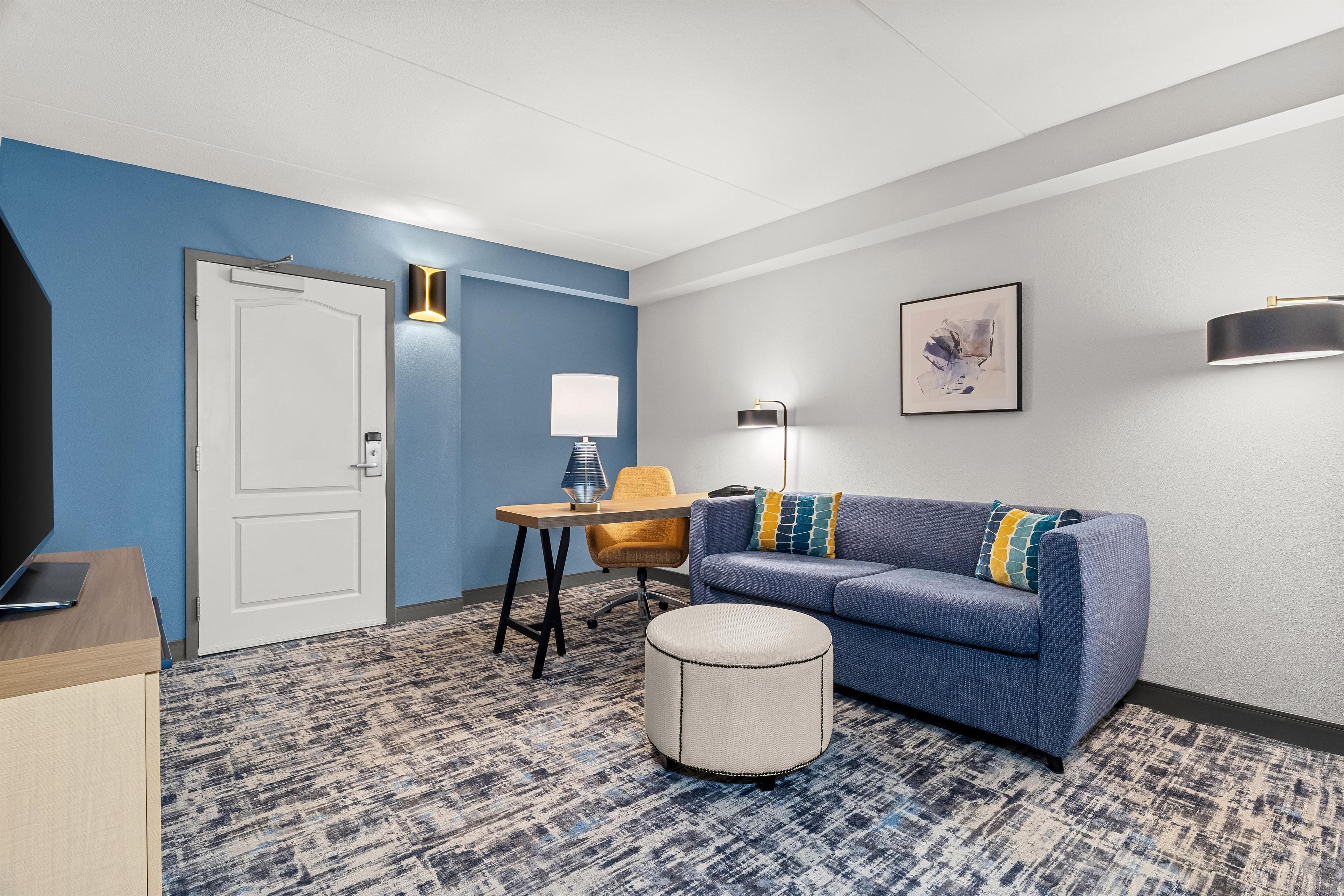 A hotel room with a blue accent wall, a sofa with patterned cushions, a small round ottoman, a desk with a lamp, and a framed artwork on the wall.