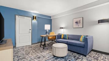 A hotel room with a blue accent wall, a sofa with patterned cushions, a small round ottoman, a desk with a lamp, and a framed artwork on the wall.