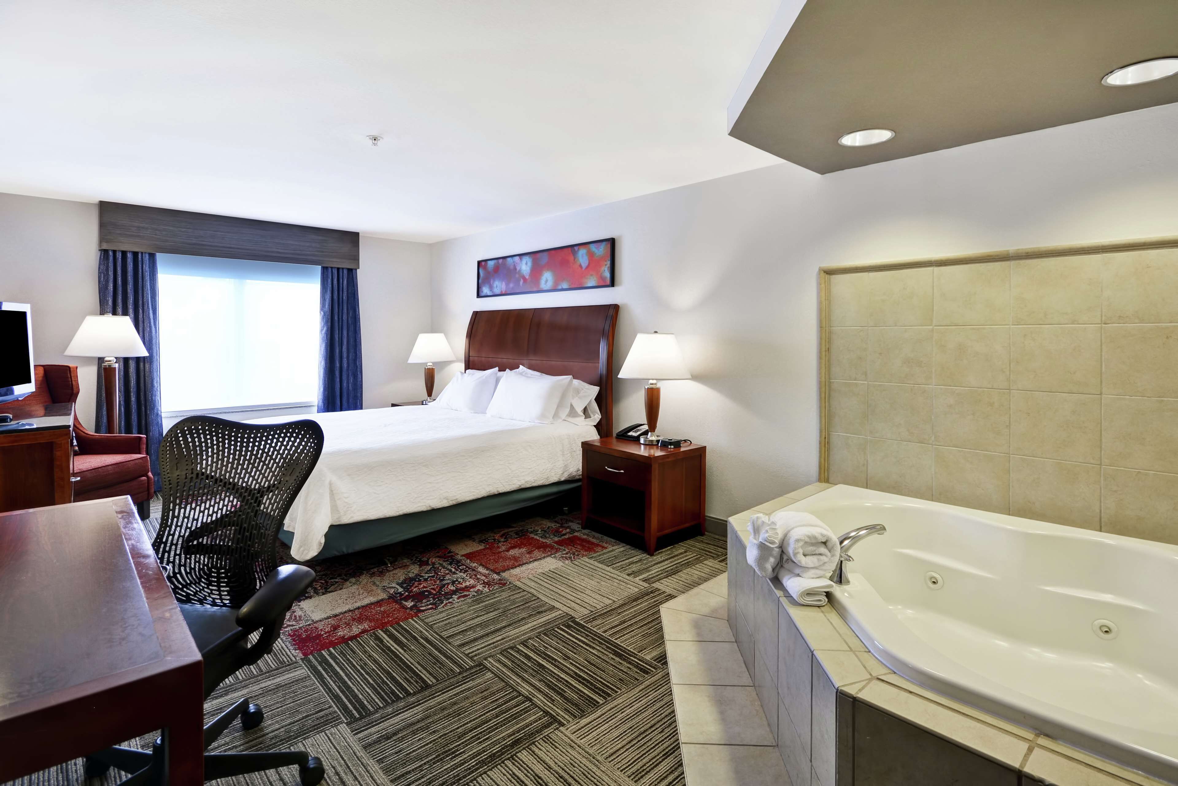 A hotel room with a bed, desk, chair, lamps, and a large bathtub.