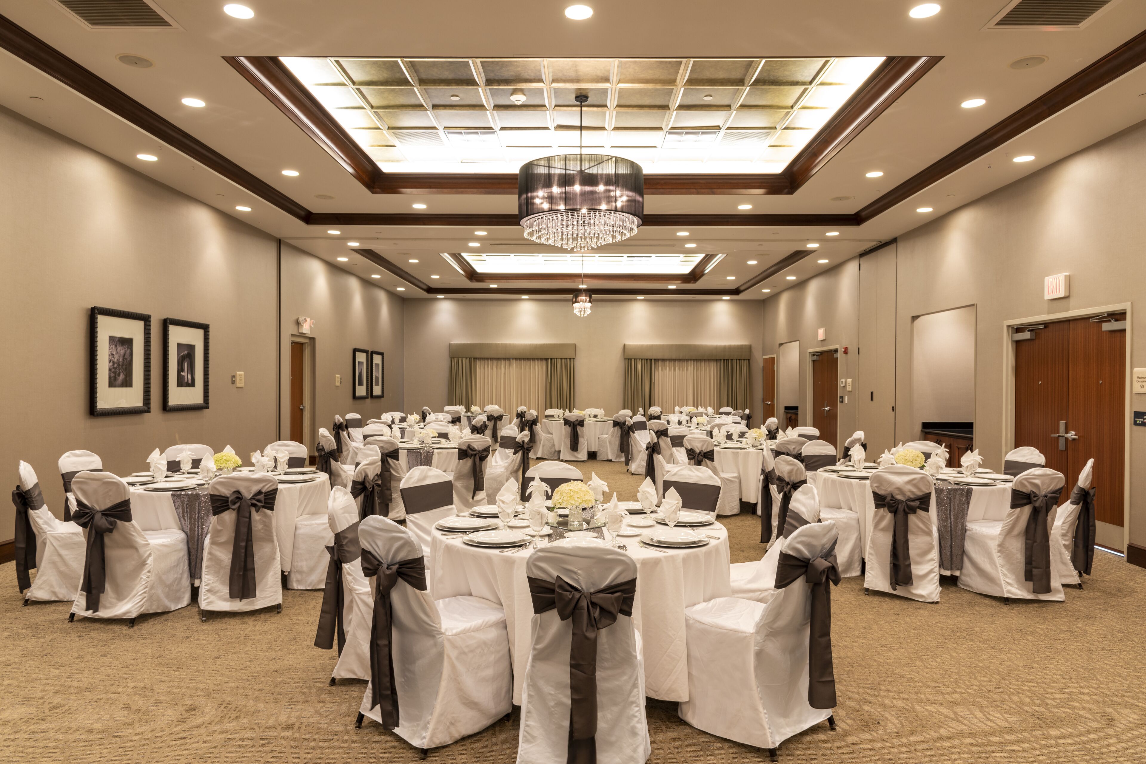 Une salle de banquet avec des tables rondes, des chaises ornées de rubans blancs et gris, un lustre et des tableaux encadrés accrochés aux murs.
