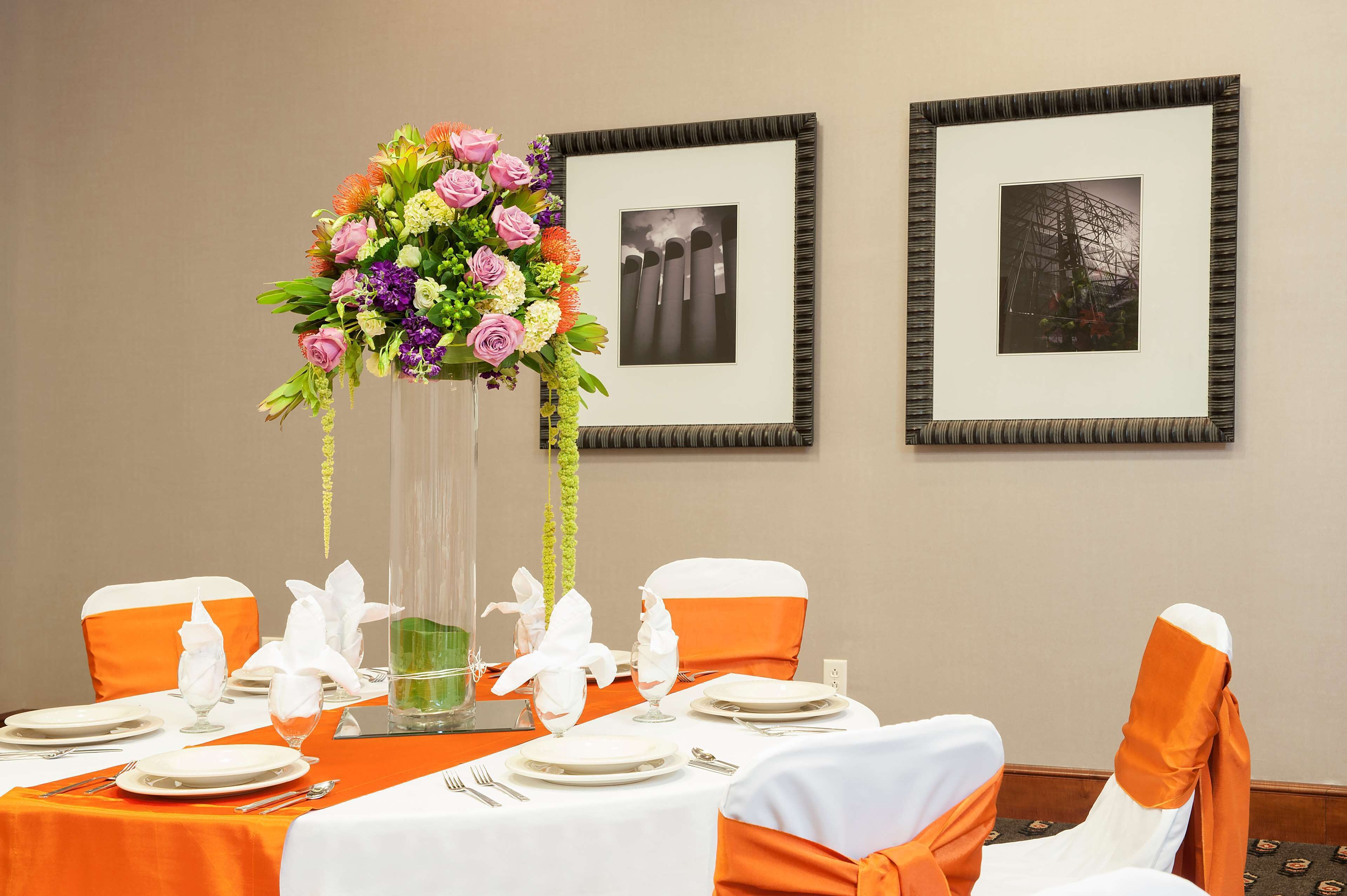 A dining table set with white plates, silverware, and orange sashes on chairs. A tall vase with flowers is centered on the table.